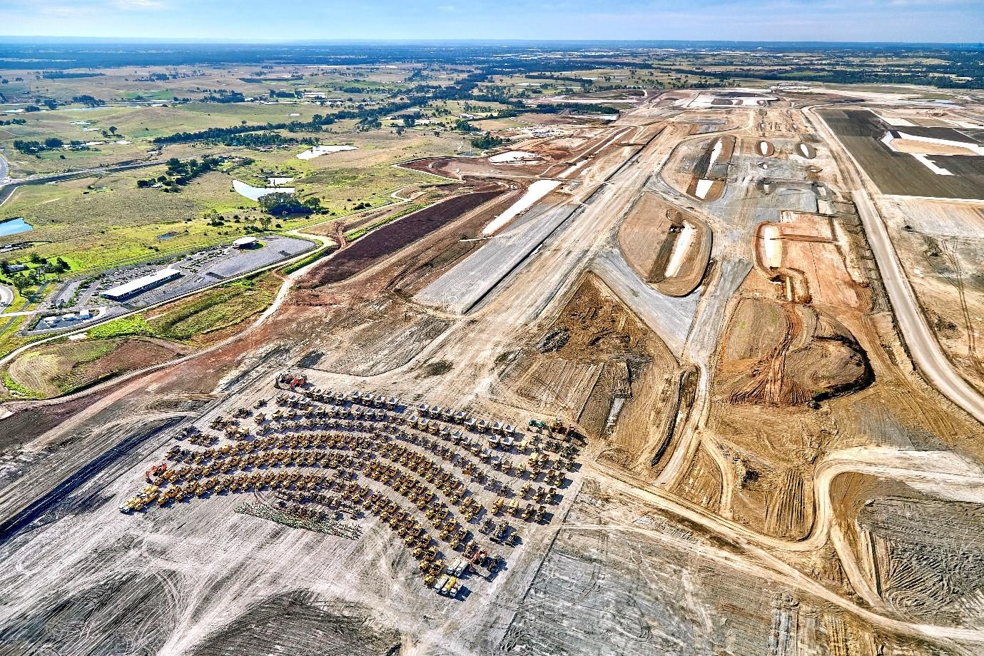 Aerial picture of plant equipment grouped together in a staged photograph celebrating the end of bulk earthworks. In the background, the outline of a runway and taxiways can be seen.